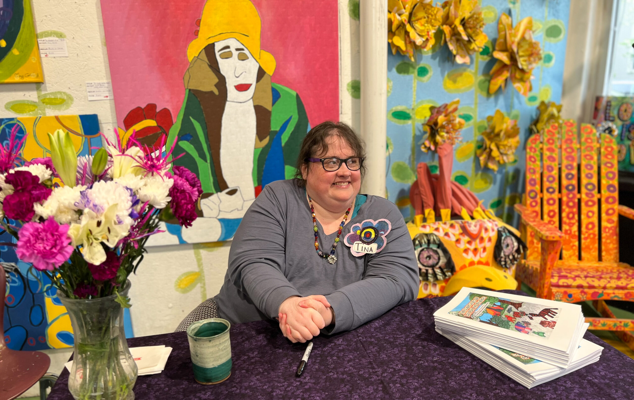 A person is seated at a table with only their upper body visible, hands clasped together, and smiling. They are wearing a name tag that reads "Tina" and are seated at a table with a stack of books. There is a vase filled with flowers off to the side and behind them is a colorful painting.