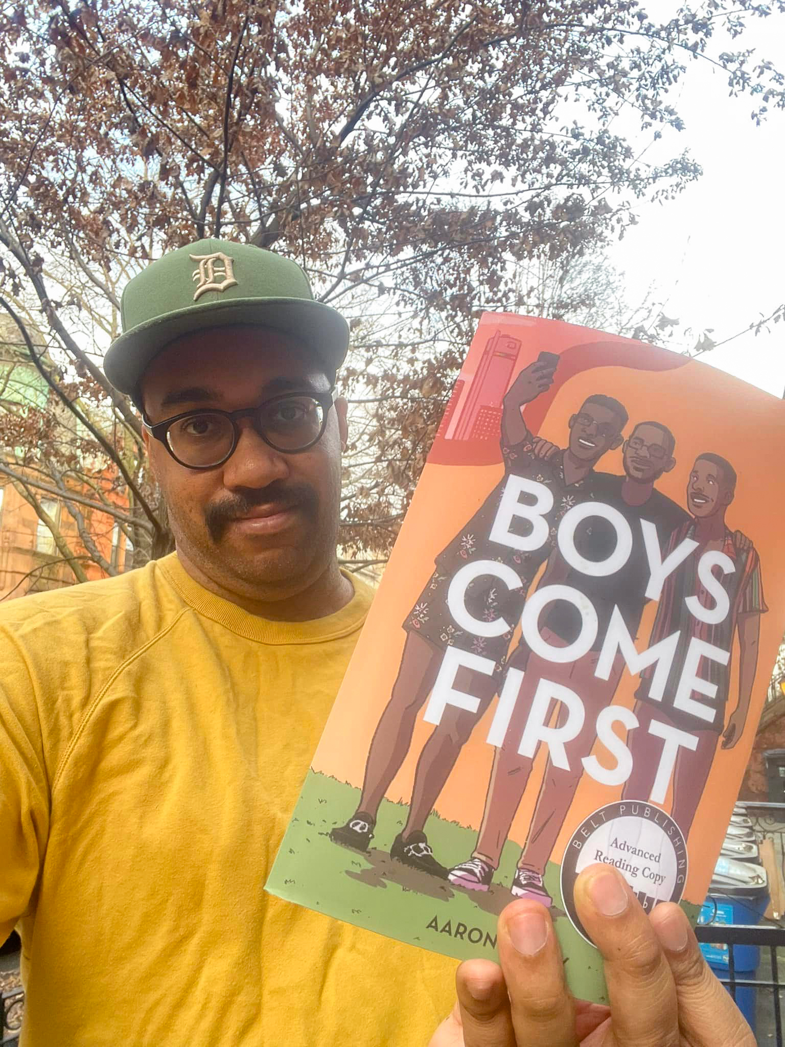 A person of dark skin tone with a mustache, wearing black-rimmed glasses and a green hat. They are wearing a yellow t-shirt and looking at the camera while holding up a book titled 'Boys Come First.' The book has an illustrated cover with three dark skin people smiling and embracing.