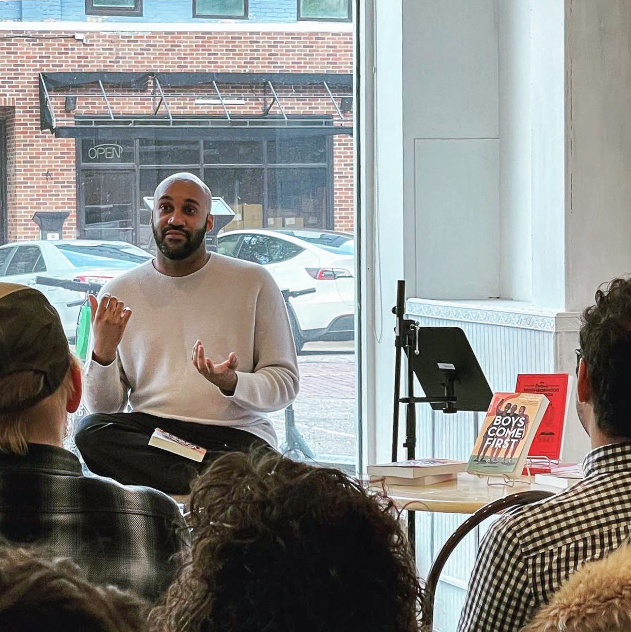 A person of dark skin tone with buzzed hair and a short beard, wearing a light grey pullover, is seated and gestures with his hands as he talks to a seated audience. There is a table beside them with a couple of books.