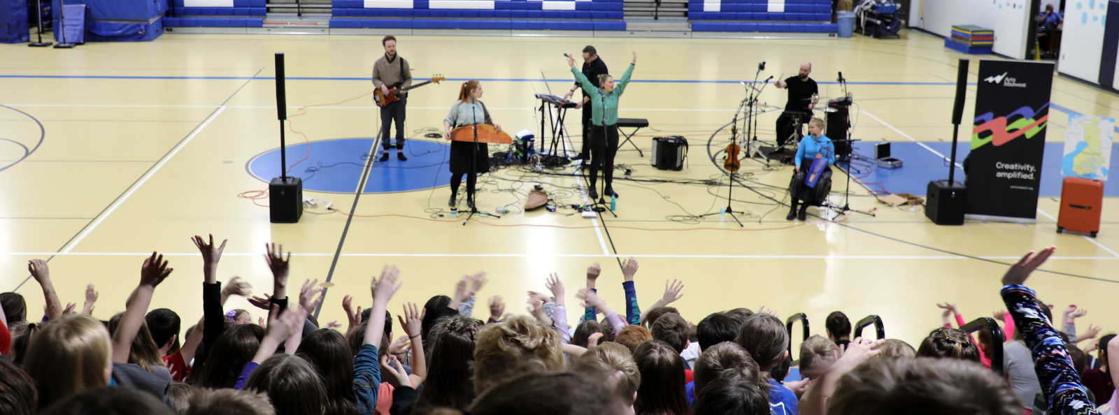 A band performs for a school of elementary students with their backs facing the camera, arms waving in the air.