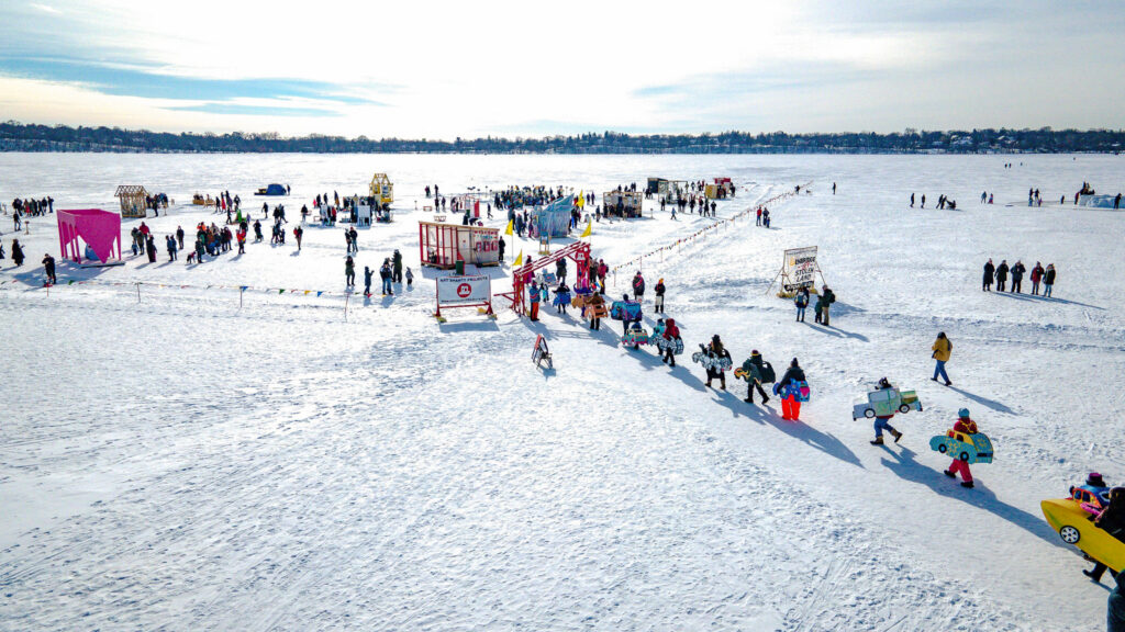 An aerial view of a frozen lake dotted with small, colorful, handmade shelters and small groups of people. There is a line of people walking towards it dressed in winter gear, holding car-shaped costumes around their hips.