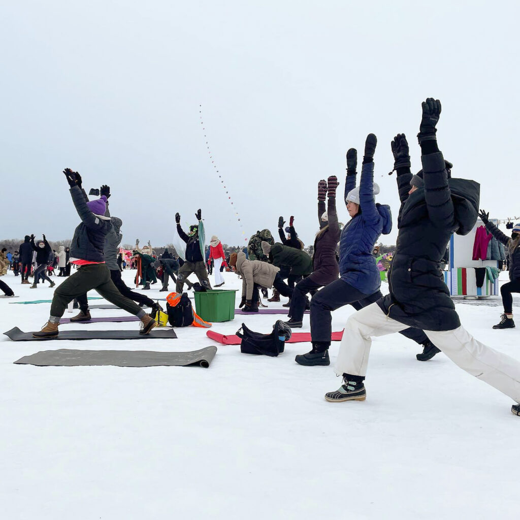 A group of people lined up, arms and legs stretched into a yoga sun pose. They are all wearing heavy winter gear as they do yoga on a frozen lake.