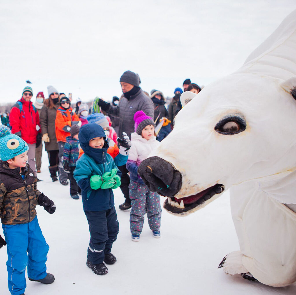 A handful of young children wearing full winter gear look at a large papier-mâché polar bear on a frozen lake. The background of the photo is dotted with other people in colorful winter gear, approaching the bear.