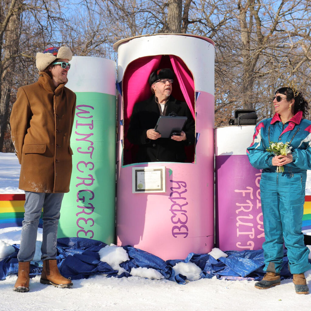 A couple stand across from each other, dressed in winter gear with the ground frozen over. They are smiling at each other as they listen to a licensed minister reading out loud from a colorful, makeshift chapel.