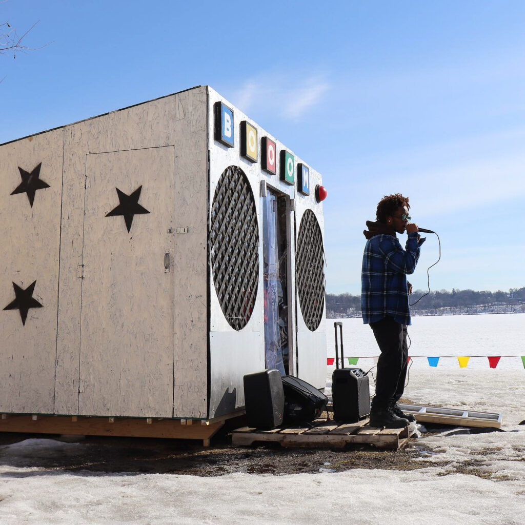 A person of dark skin tone holding a microphone performs in front of a makeshift shelter. The shelter is an interactive art installation that looks like a large boombox. It is situated on a frozen lake.