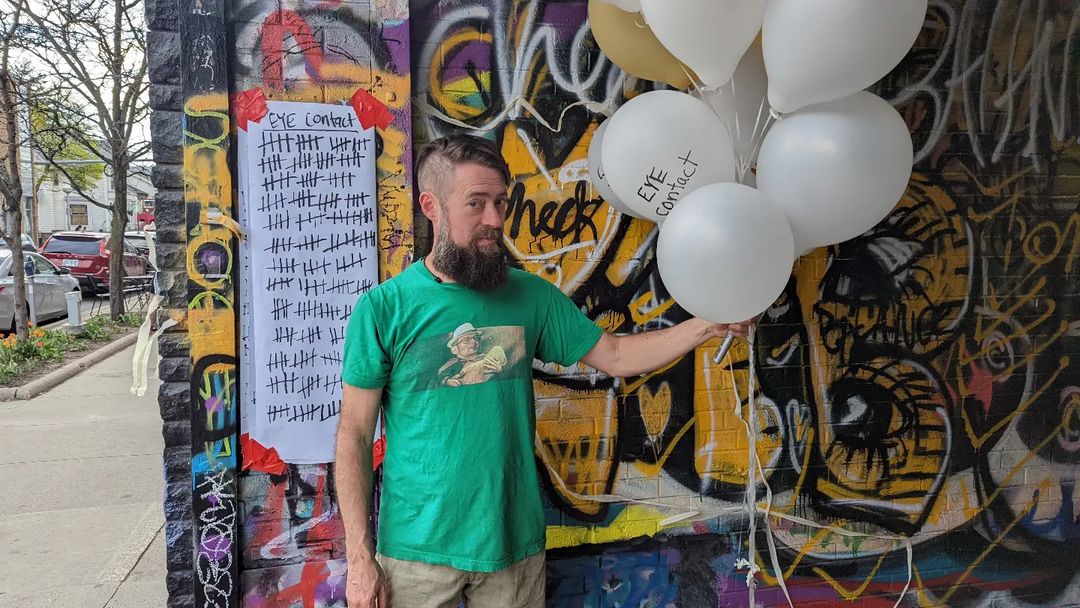A man holding a bunch of white balloons stands against a background of graffiti and a paper that says eye contact and has tally marks.