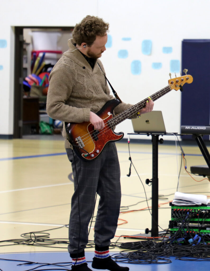 A man plays bass guitar in a school gymnasium