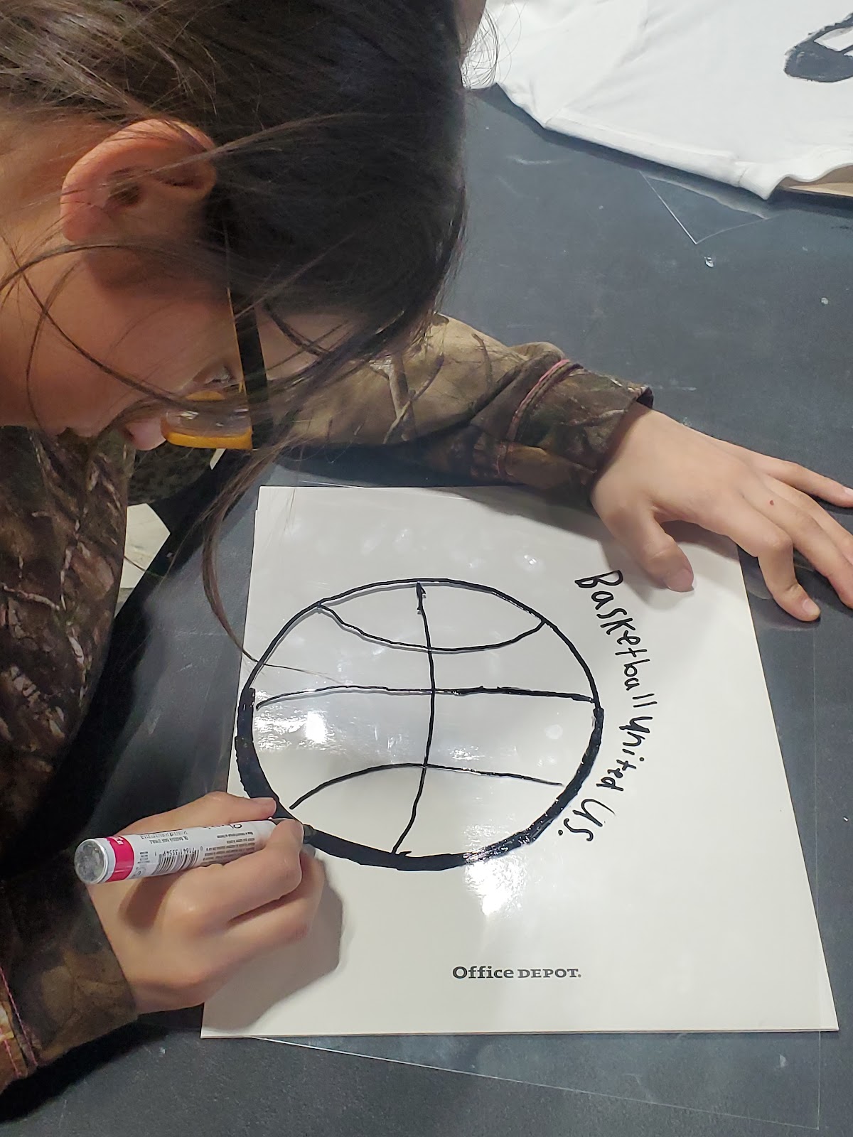 A young student traces a basketball and text that reads "Basketball Unites Us" onto a piece of paper