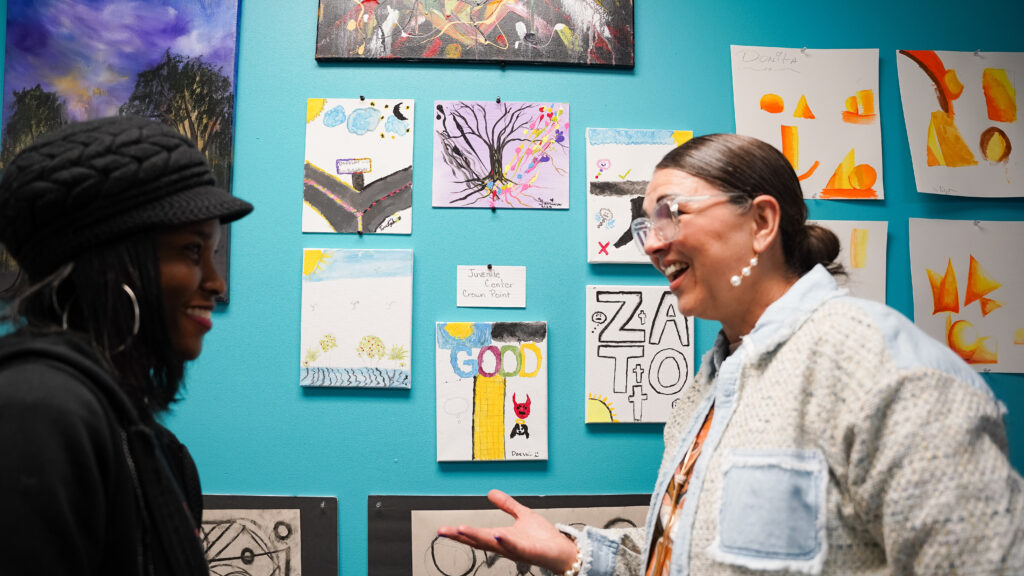 Two women talk enthusiastically in front of a teal wall covered in colorful paintings and drawings.
