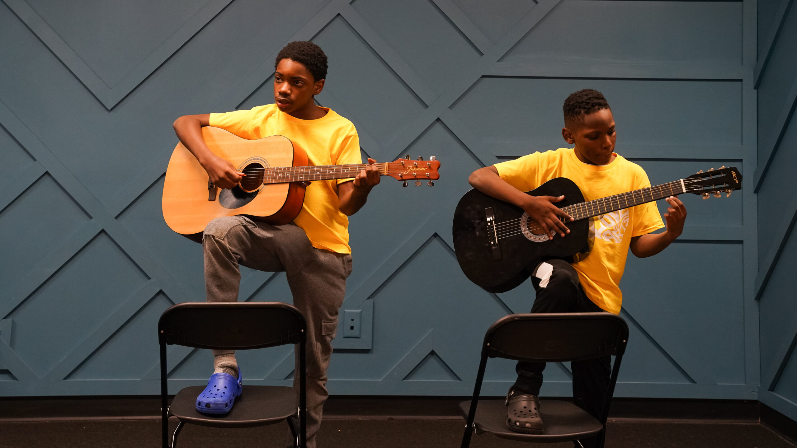 Two young boys hold guitars, with their feet up on chairs as they play