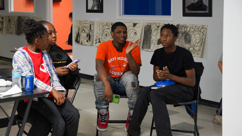 Four young people sit on chairs and talk in an art gallery space. Daveed Holmes