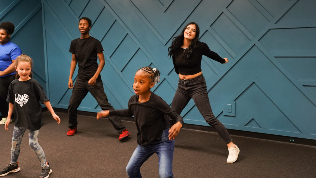 Students of several ages participate in a dance class with smiles on their faces.