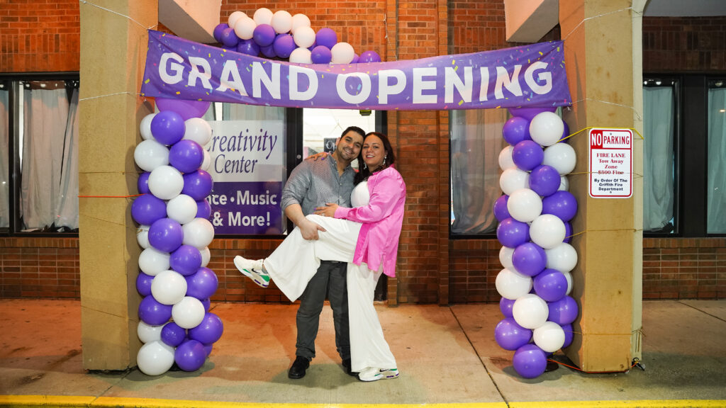 Two people smile as they stand under a vibrant balloon arch and signs that read Grant Opening Creativity Center Dance Music and More.