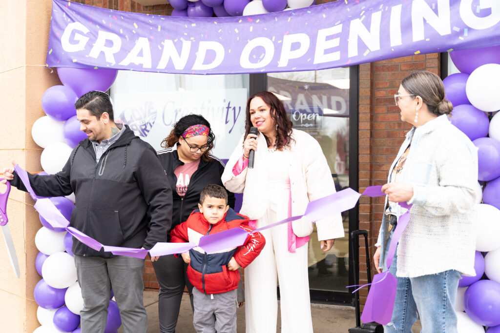Two adults hold up a ribbon for cutting underneath a balloon arch and a sign that reads Grand Opening. A woman smiles as she speaks in to a mic in the middle.