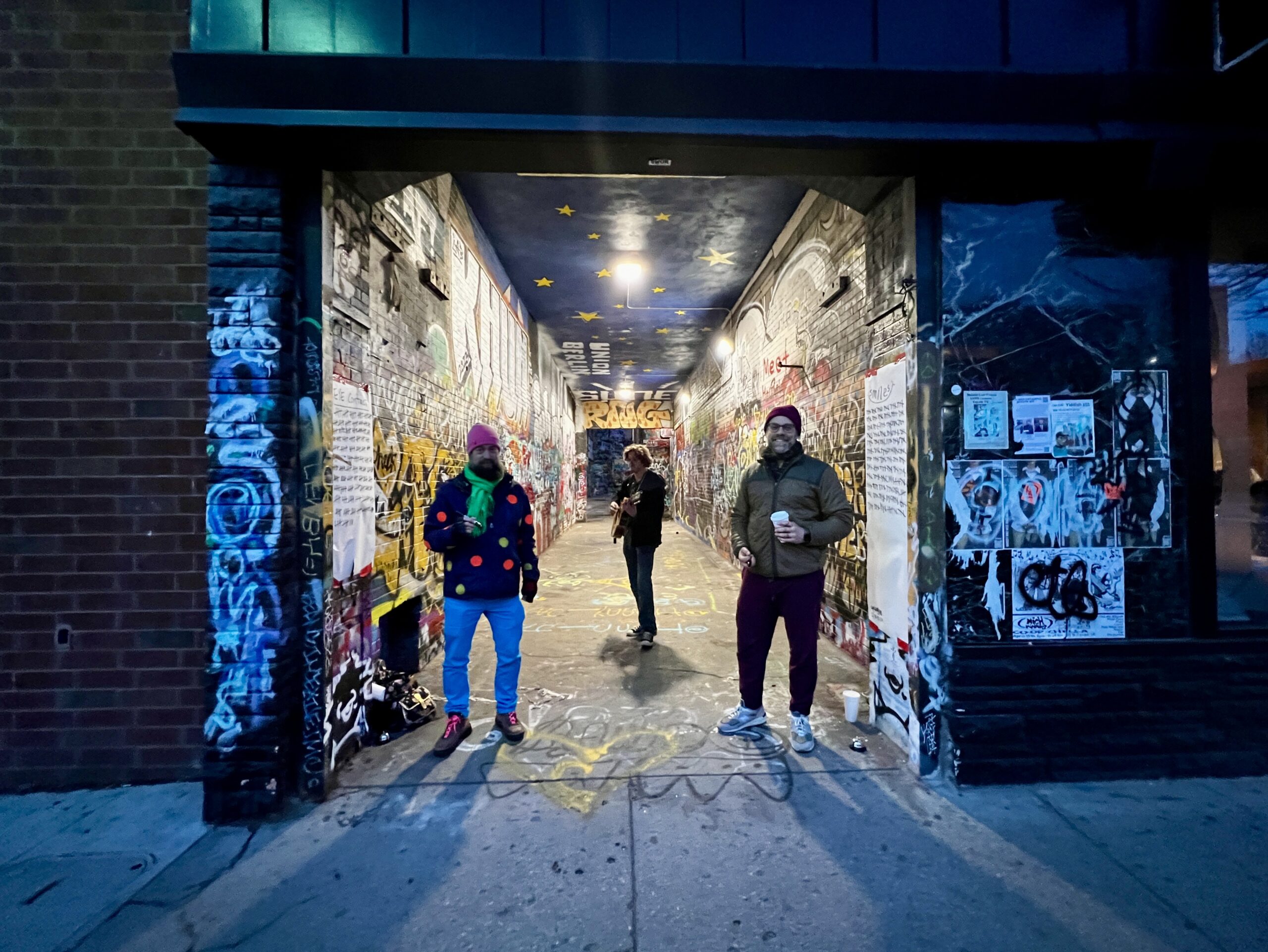 Three men stand in an alley full of graffiti at night, with two pieces of paper, one on either side of the alleyway.