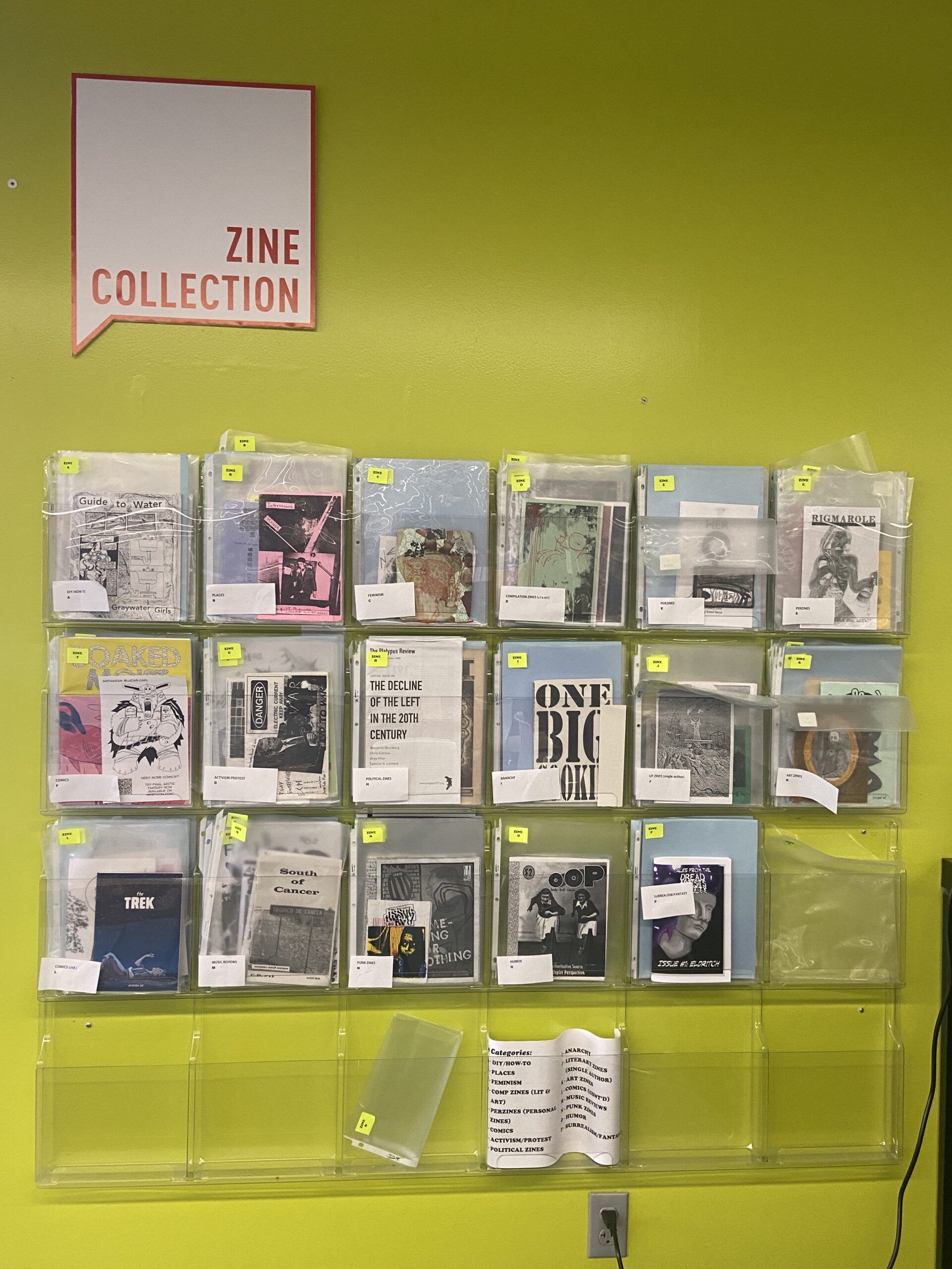 On a bright chartreuse-colored wall, there are over two dozen plastic magazine holders mounted and filled with zines. There is a zine above it that reads "Zine Collection." Several of the magazine holders at the bottom are empty.