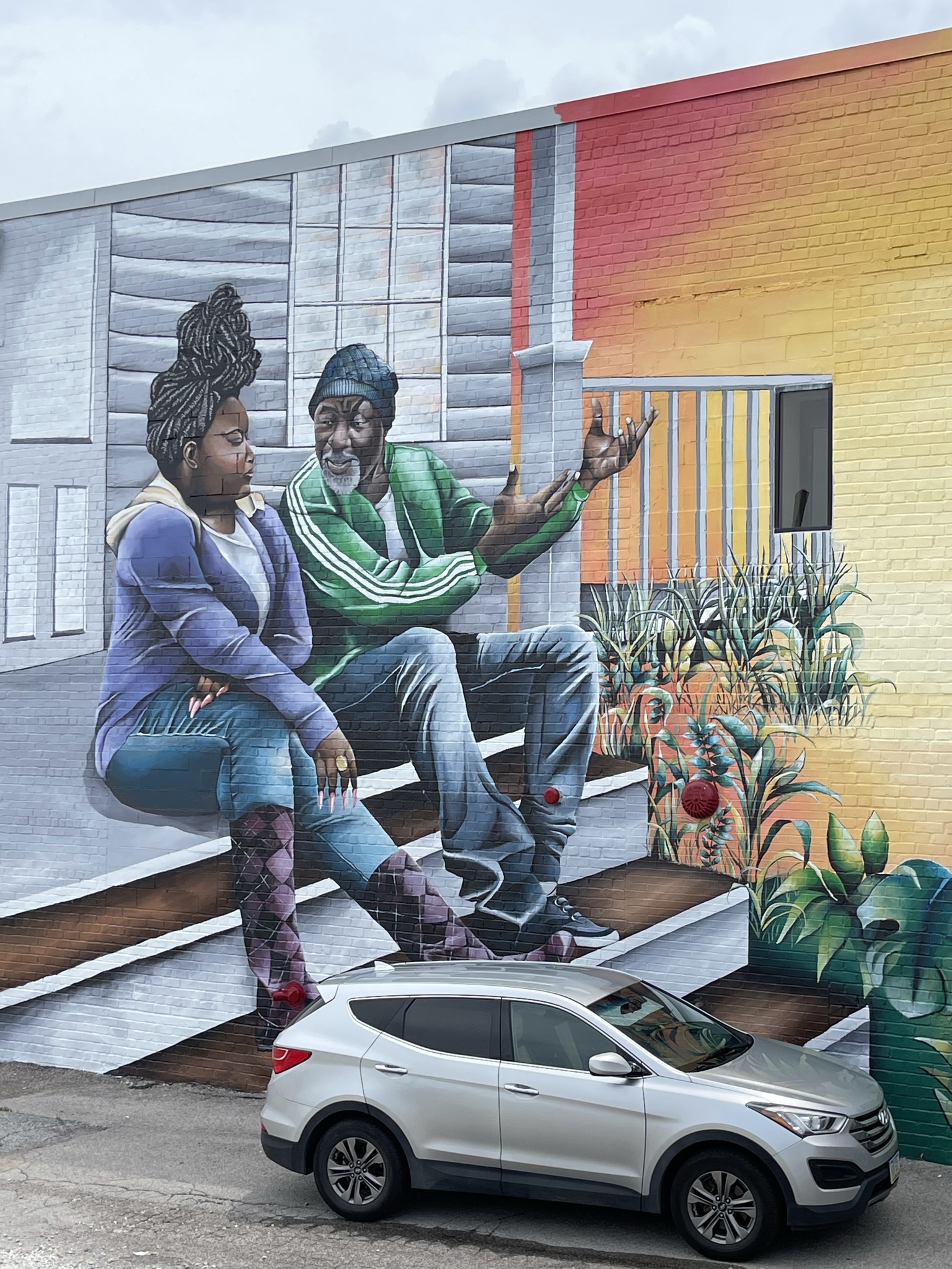 A mural depicting two people of dark skin tone sitting on the porch of a white house. The older person depicted in the mural is gesturing with their hands, while the other listens. There are tones of yellow and red, almost a sunset, in the background with some plants.