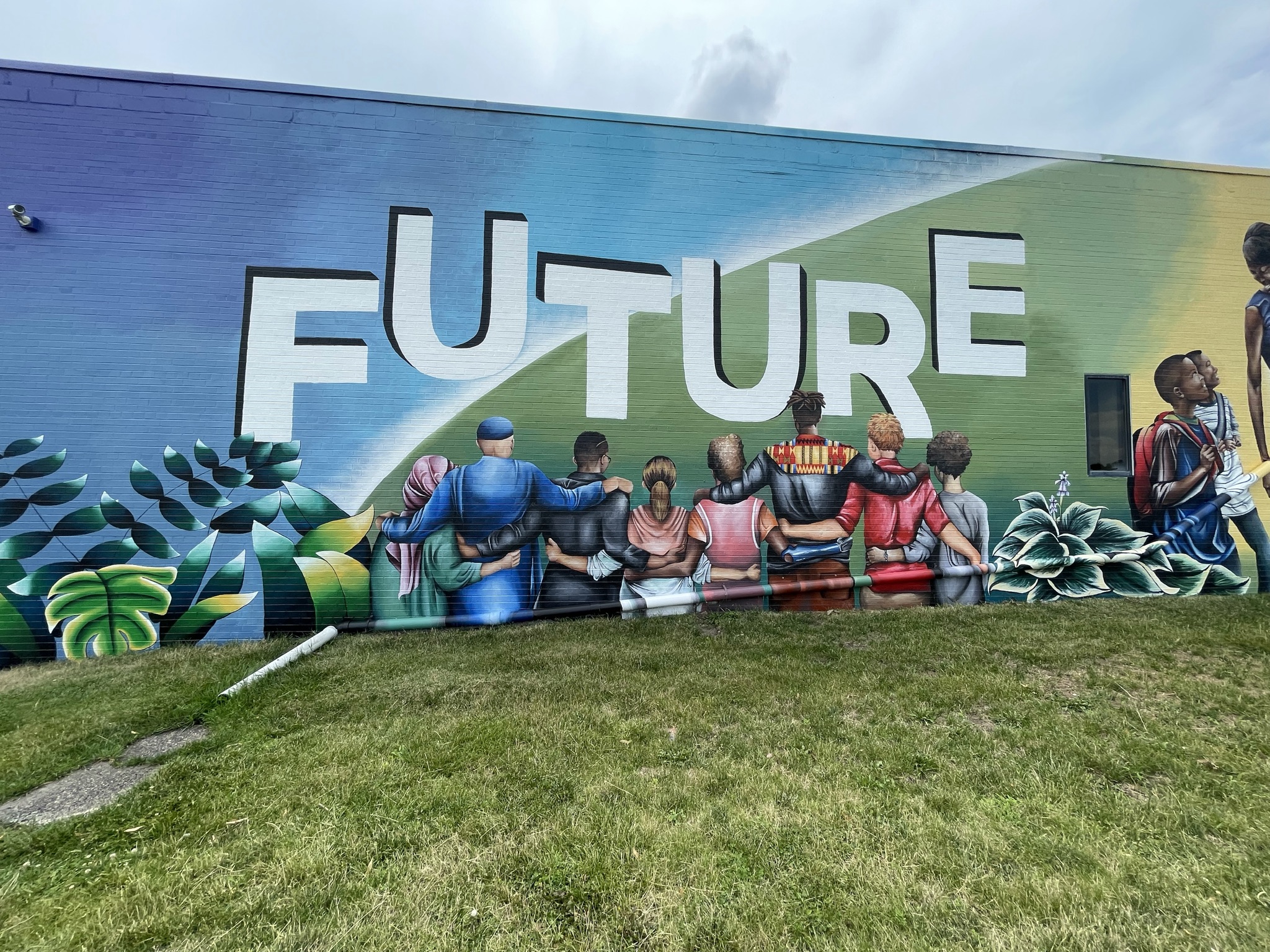 A mural depicting a group of people with dark skin tones and dark hair embracing with their backs to the viewer. They are placed under letters that read "future" in all caps across a blue and green horizon-like background.