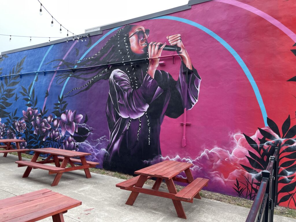 A mural depicting a young Black woman, wearing braids and baby hair, singing into a microphone with one fist in the air. They command attention amid flowers, black butterflies. The background of the mural is a color face from blues, to purples, and pinks. There are a handful of brown picnic tables by the mural wall.