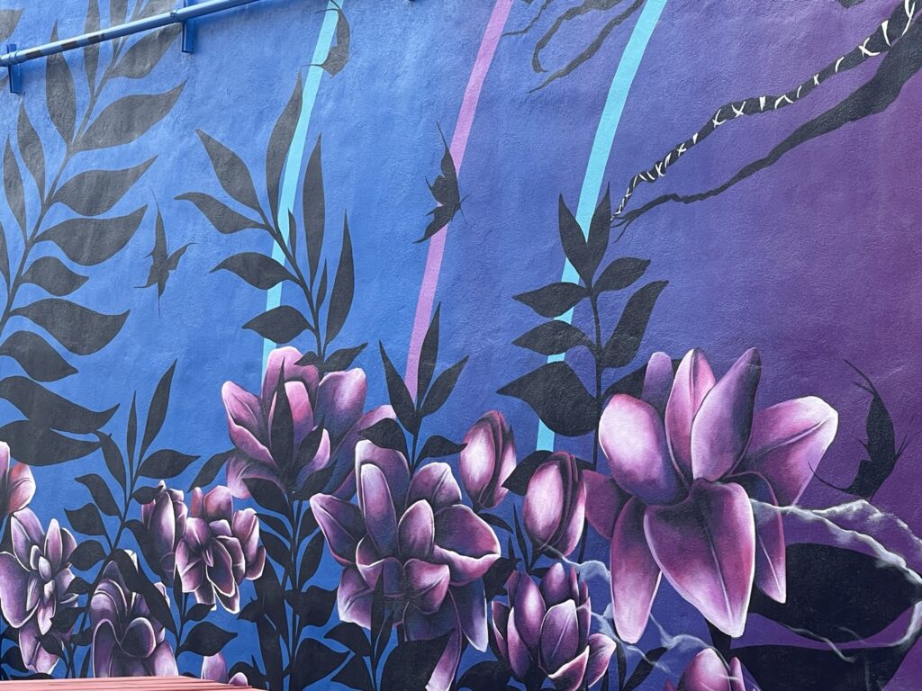 A close-up of a mural depicting moody lotus flowers in pink and purple tones, with black butterflies flying above them and dark foliage. All of this is on top of a color-fade background of blue and purple tones.