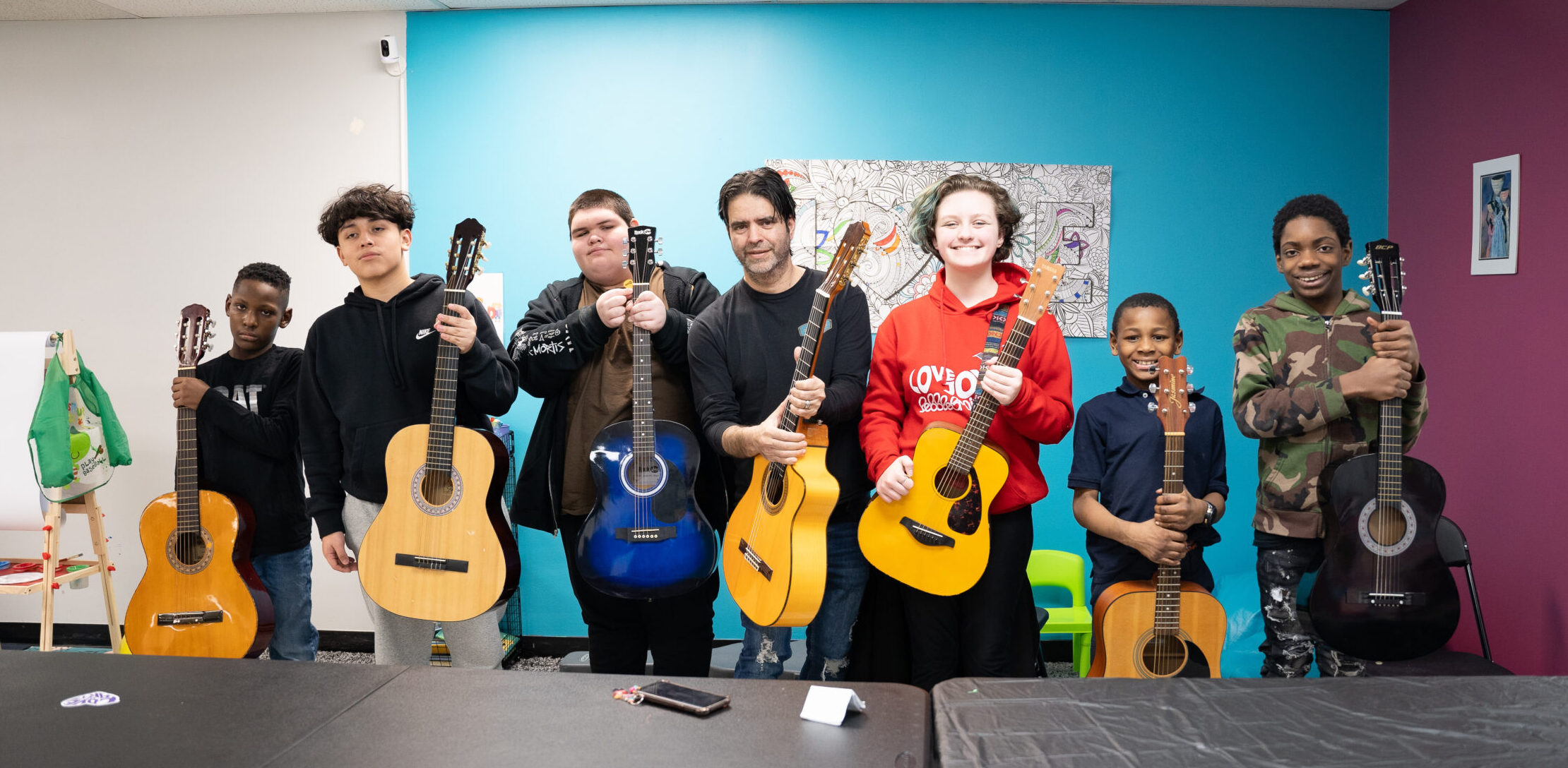 Six students and one instructor hold their guitars up to the camera and smile
