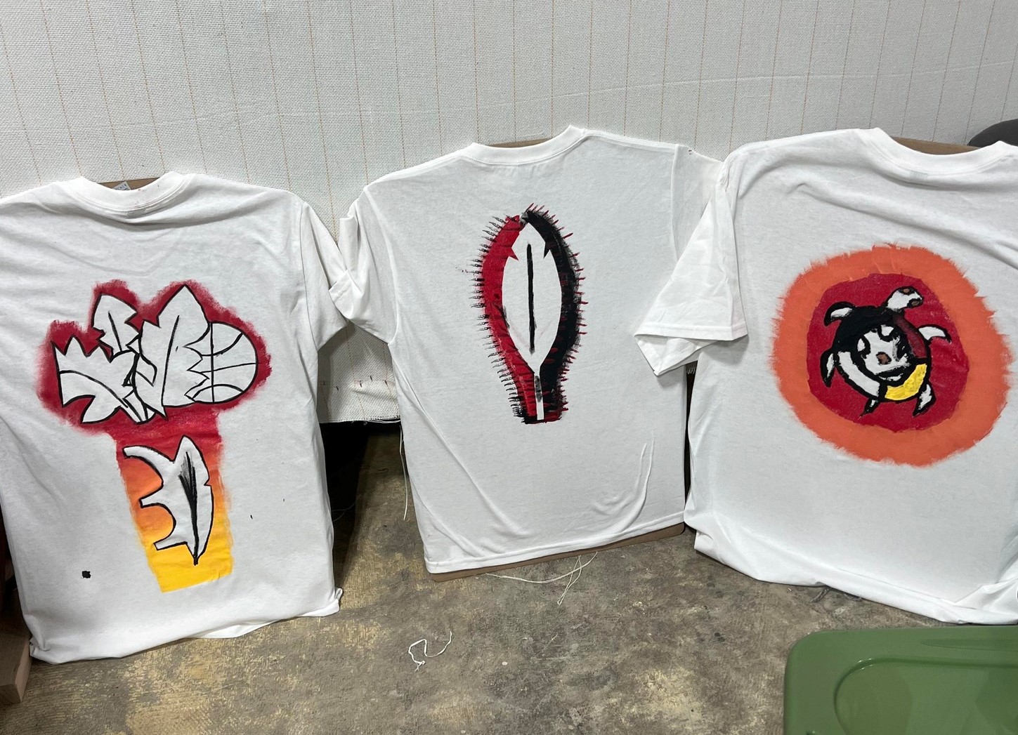 Three t-shirts with designs including a feather, Turtle Island, and leaves and basketballs.