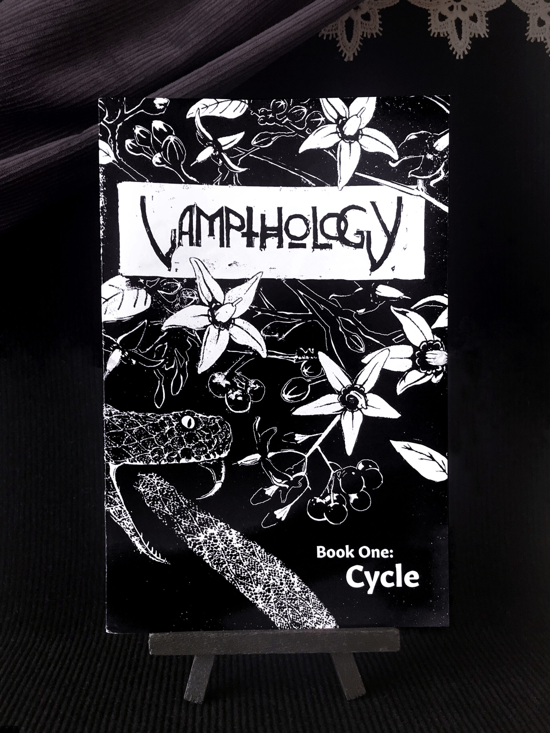 A black and white zine cover depicting a snake about to bite its tail, surrounded by flowers and other foliage. The cover has lettering that reads 'Vampthology' at the top center, and 'Book One: Cycle' in the bottom right-hand corner.