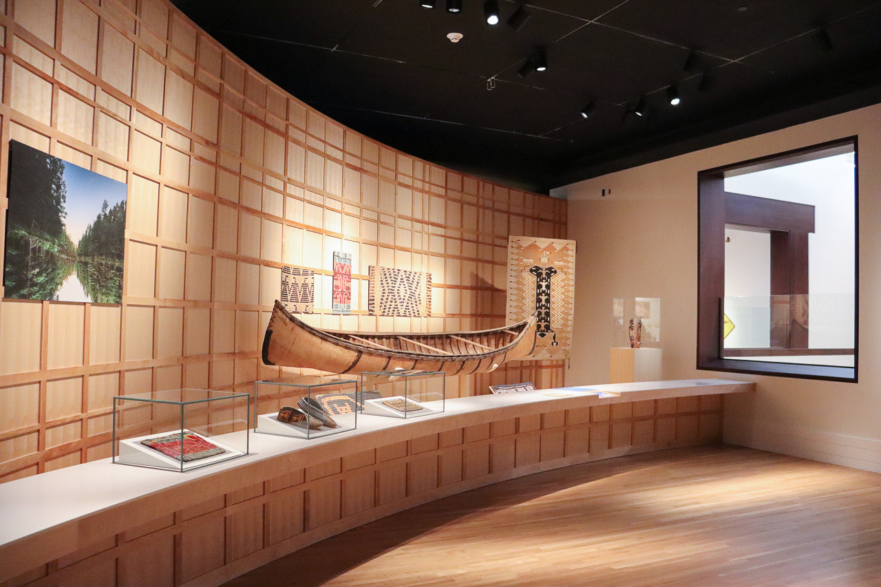 A wide view of a museum gallery space with photography, woven artworks, and a canoe on display.