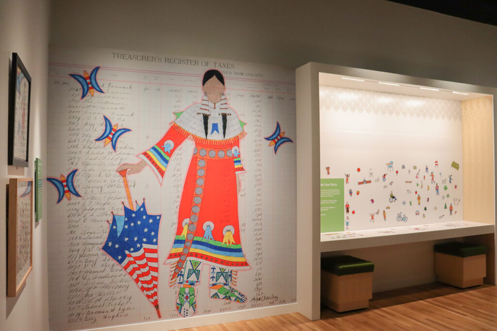 A large artwork on the wall of a museum gallery depicts a person in traditional Native American regalia. The drawing is on ledger paper.