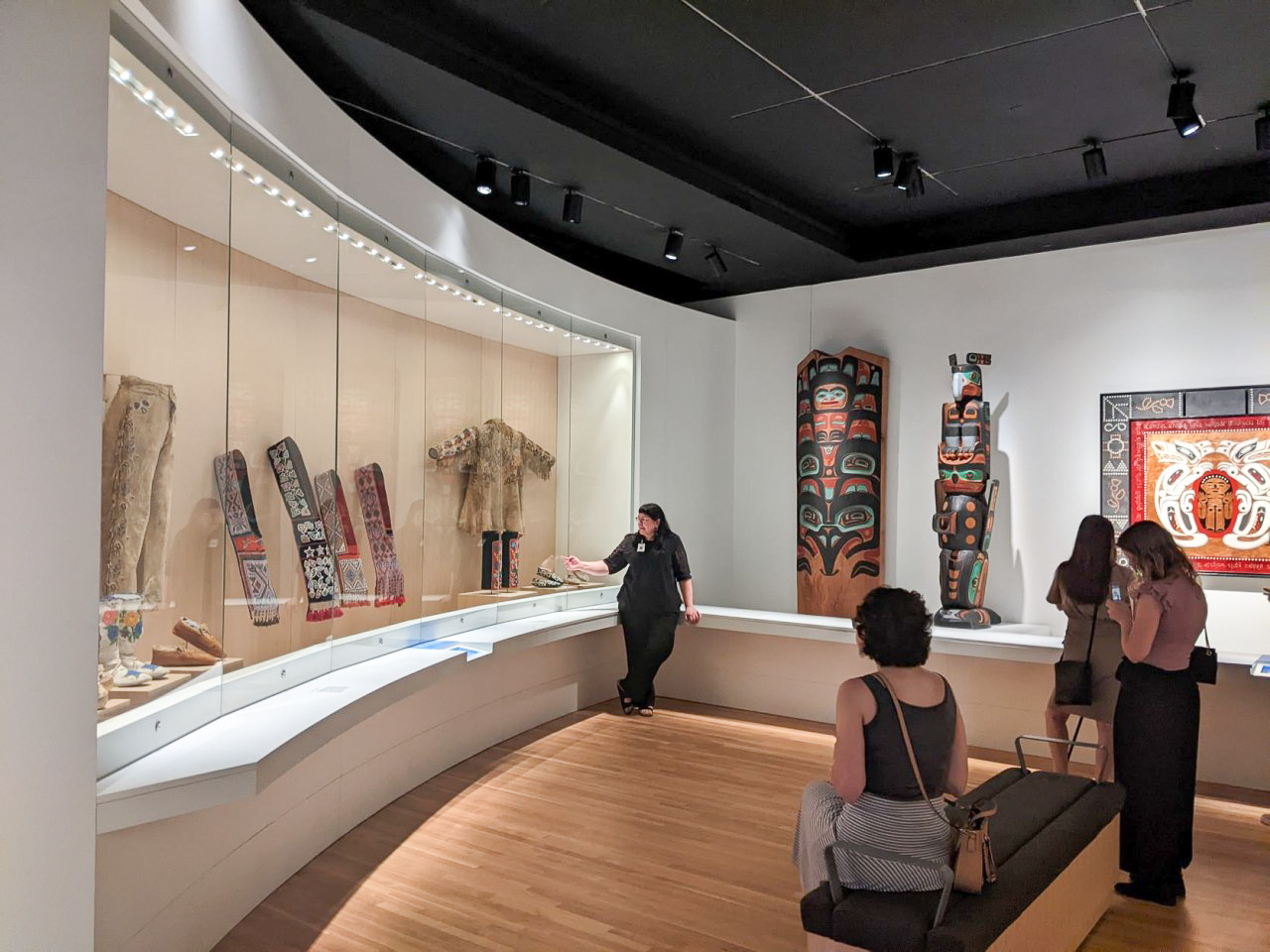 A wide view of a museum gallery space with three people listening to the curator talk about the exhibit. The exhibition is of Native American and Indigenous Art.