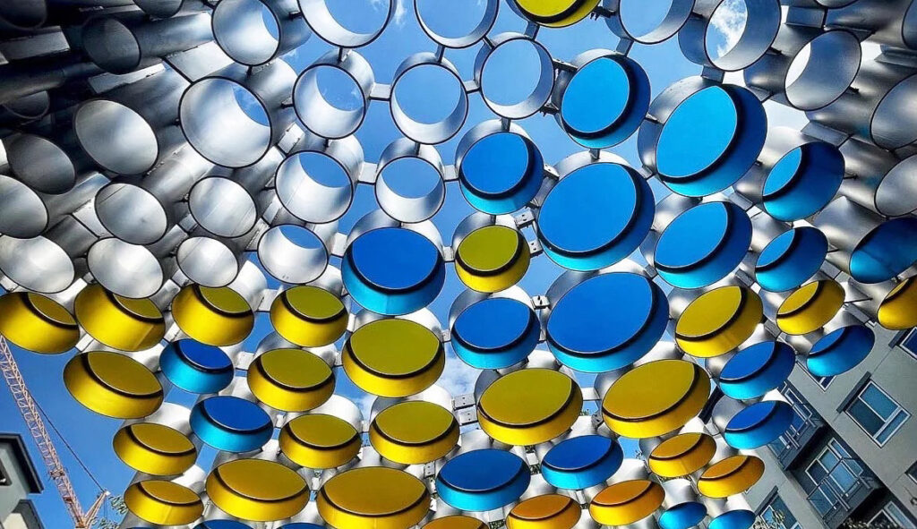 A web of circular metal rings, in silver, blue and yellow, of varying sizes make up this public artwork. It acts as a shade canopy. The photograph is captured from the ground up, with blue skies as the backdrop.