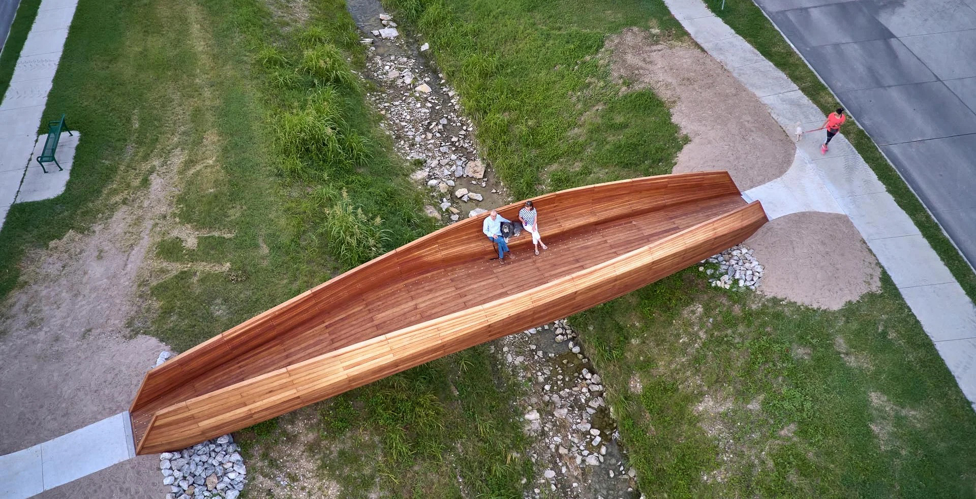 An aerial view of a public artwork made of cedar wood. It is a short bridge structure that extends over a small rocky creek. The piece almost looks like an oversized canoe. There are two people with a dog sitting on a seating that extends from the inside of the artwork.