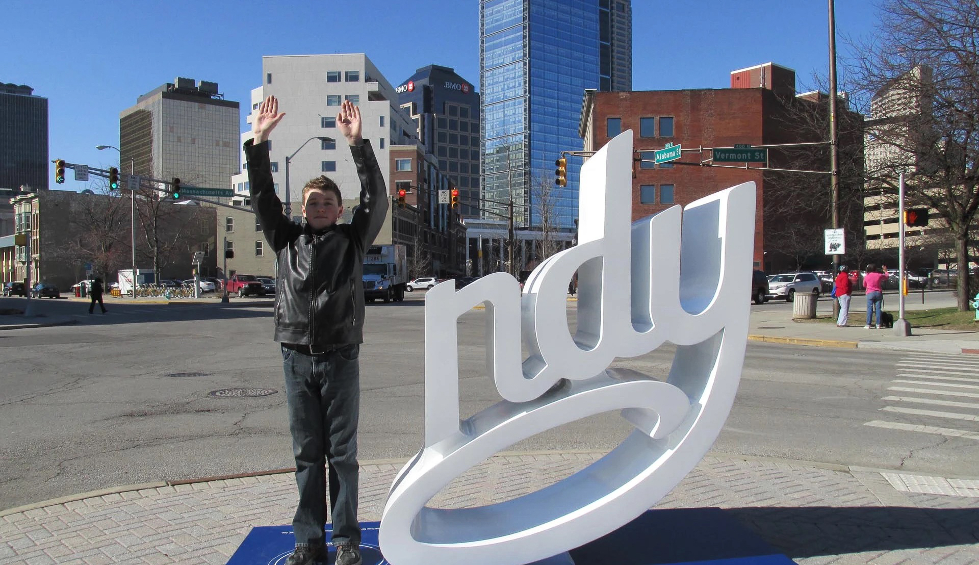A young person of light skin tone in dark clothing raises both their hands up in the air as they stand next to a public artwork that spells 'ndy' in lower case cursive lettering. The person stands in for the letter 'I' to spell out 'Indy' for Indianapolis, where the public art exists.