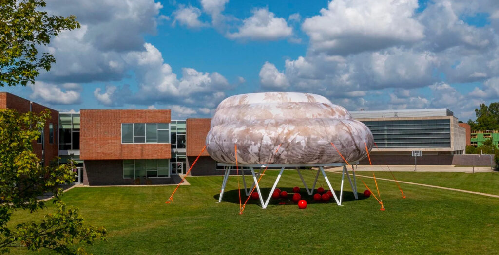 A public art installation on the exterior yard of a middle school. The artwork is comprised of a blow up structure that looks like a cloud. It is placed on a white metal structure and held down by bright orange straps. Under this, there are a number of large, bright red balls meant to be seating for people who interact with the artwork.