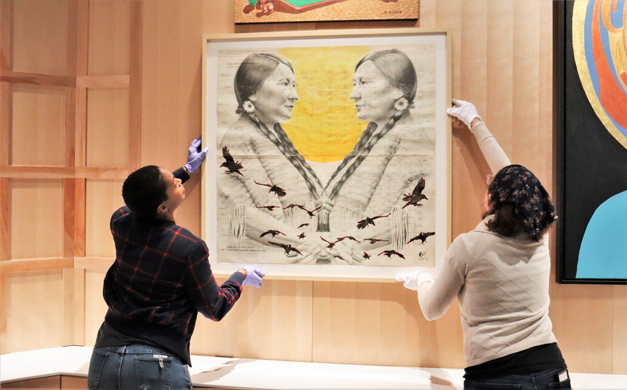 Two people hold up an artwork as they hang an exhibit in a museum gallery space. The artwork depicts two Native American people, drawn in graphite, facing each other against a bright yellow background.