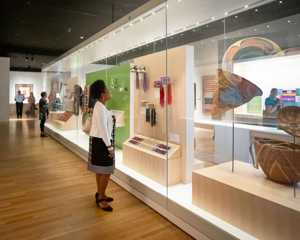 A person of dark skin tone looks at an exhibit display encased in a large plexiglass case. The pieces in the display are a wide range of beadwork and basketry.