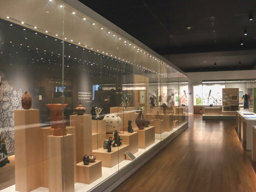 A wide, long view of a museum gallery space. Behind a large plexi exhibit area, there are over a dozen wooden pedestals of varying size displaying a diverse selection of pottery and ceramic works.