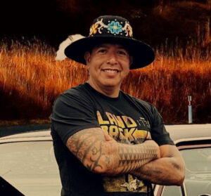 A person of medium skin tone wearing a decorative hat and a tshirt that reads Land Back stands in front of a car.