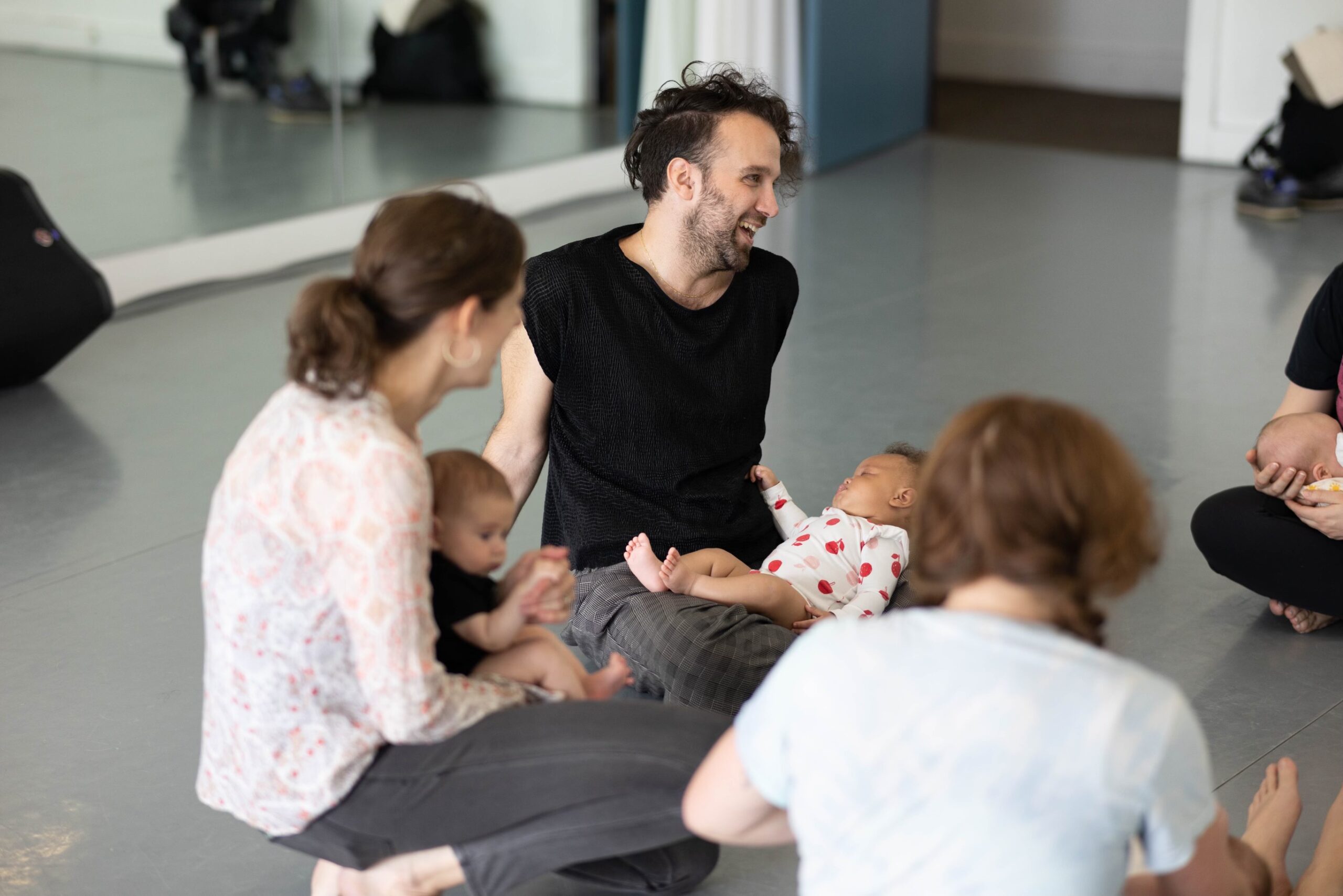 Four people of light skin tone sitting in a dance studio with babies in their laps.