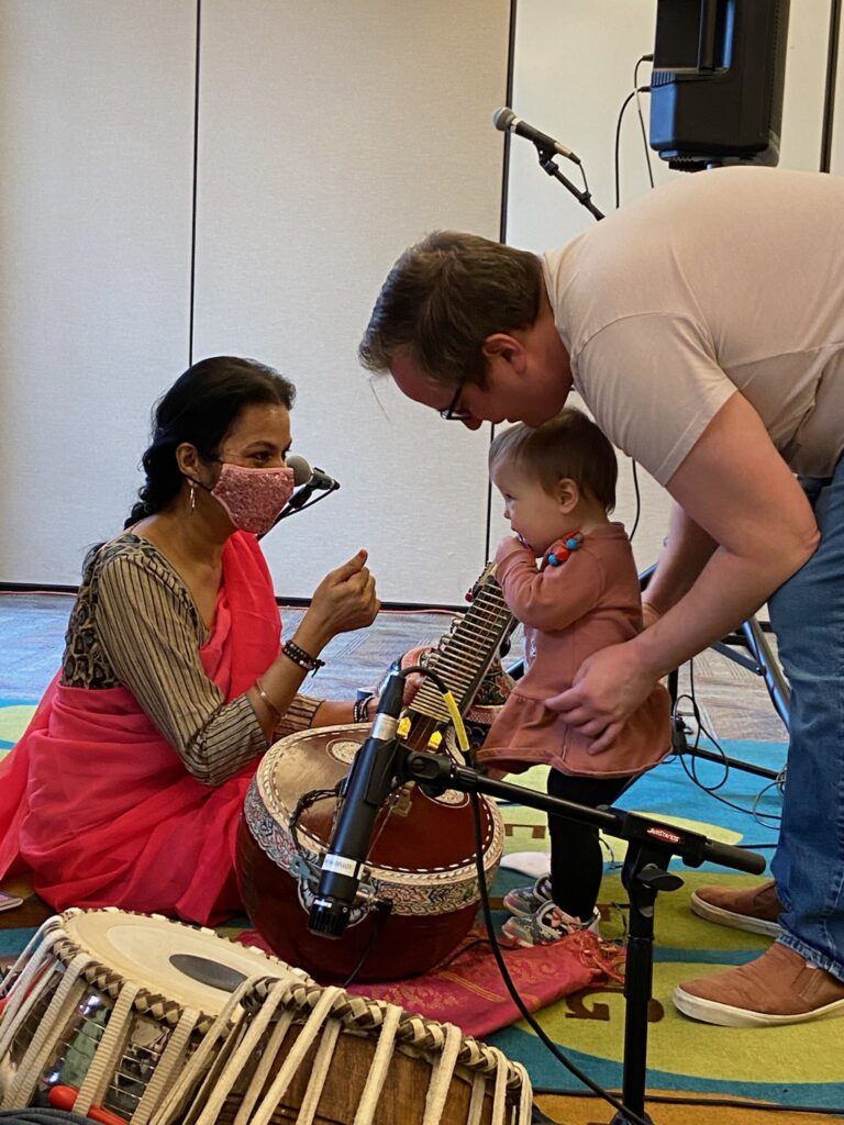 A person wearing a mask kneels by a sitar and speaks to a parent and child.