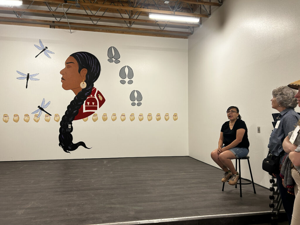 A woman with medium skin tone sits on a stool in front of a mural of a Native woman and dragonflies.