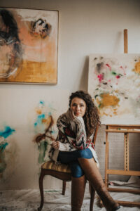 A woman with curly hair and a light skin tone poses in a studio filled with abstract paintings