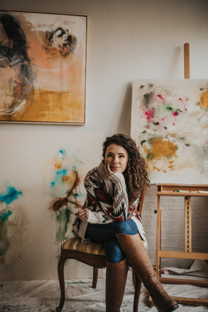 A woman with curly hair and a light skin tone poses in a studio filled with abstract paintings