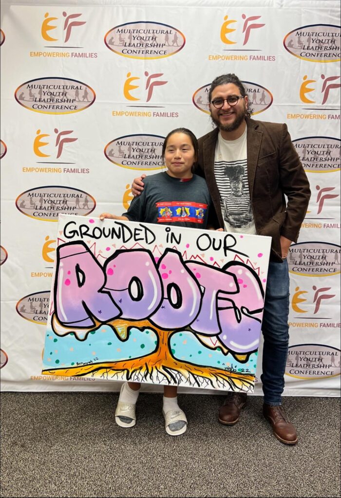 A man and a young person smile in front of a backdrop while holding a large image that says Grounded in our Roots