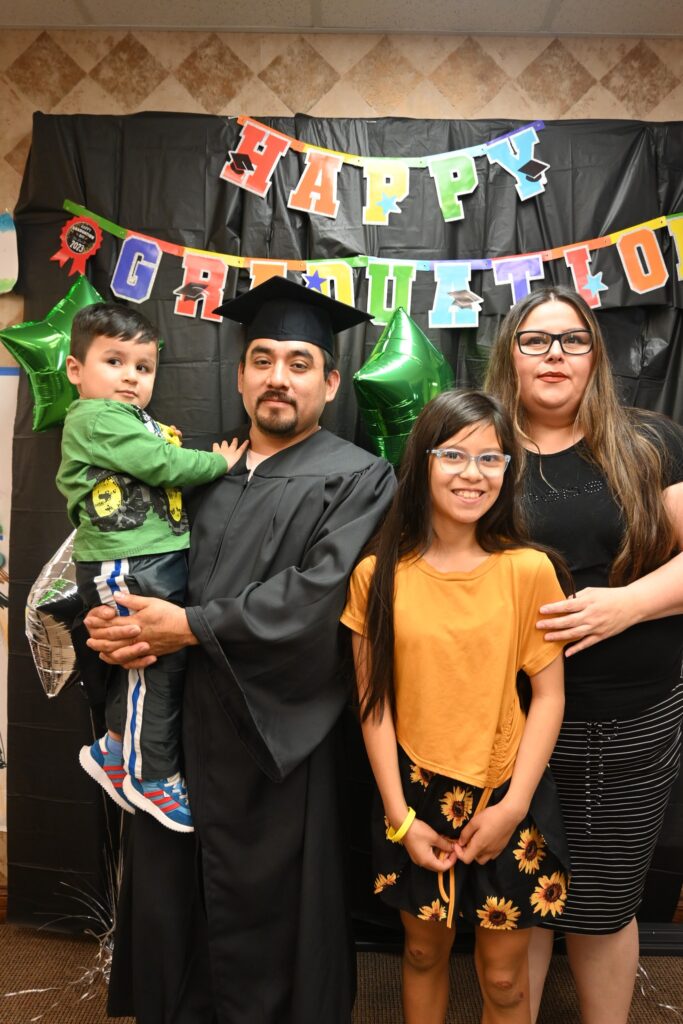 A man in a graduation robe and hat holds a young child and smiles joined by another child and a woman