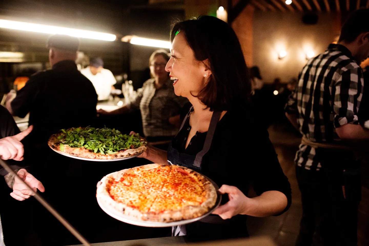 A person of medium-light skin tone holding two pizzas in a crowded restaurant.