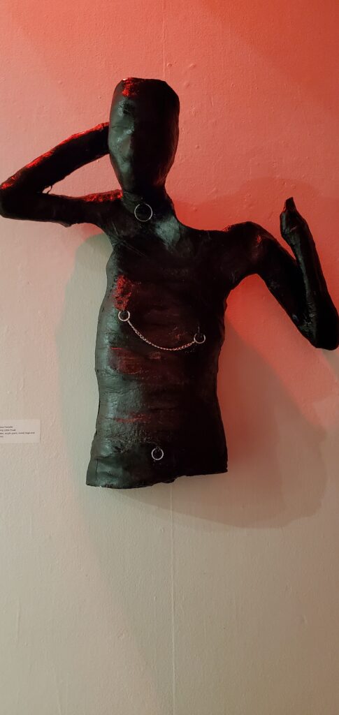 A sculpture of the top half of a person's torso painted in all black with nipple rings, a belly button ring, and a collar hanging on a wall.