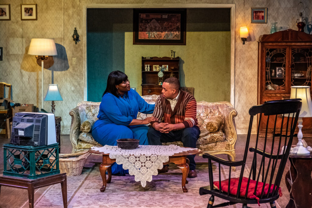 Two actors with medium to dark skin tones sit on a couch during a theatre performance. The stage is set up as a living room. One of them is holding the other softly, almost comforting them, as they face each other in conversation.