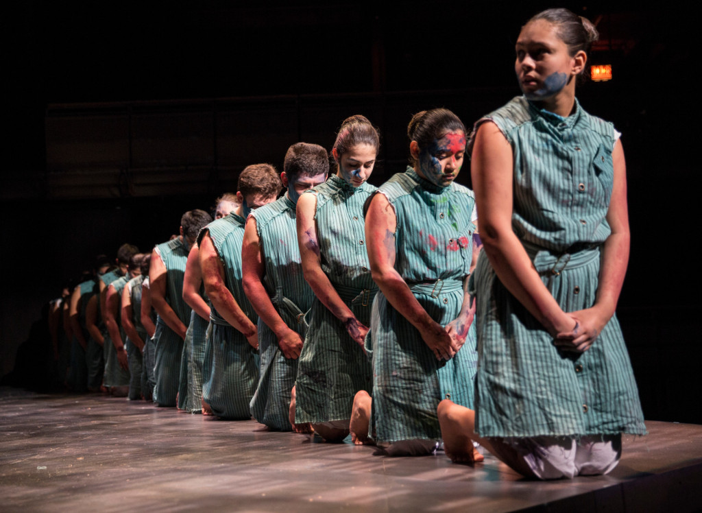 A row of young actors kneel during a theater performance. They are wearing similar teal-colored uniform. The first person has their face turned to the left, while others behind them have their heads down. Their hands are clasped in front of them, and their clothing and bodies have splotches of colorful paint.