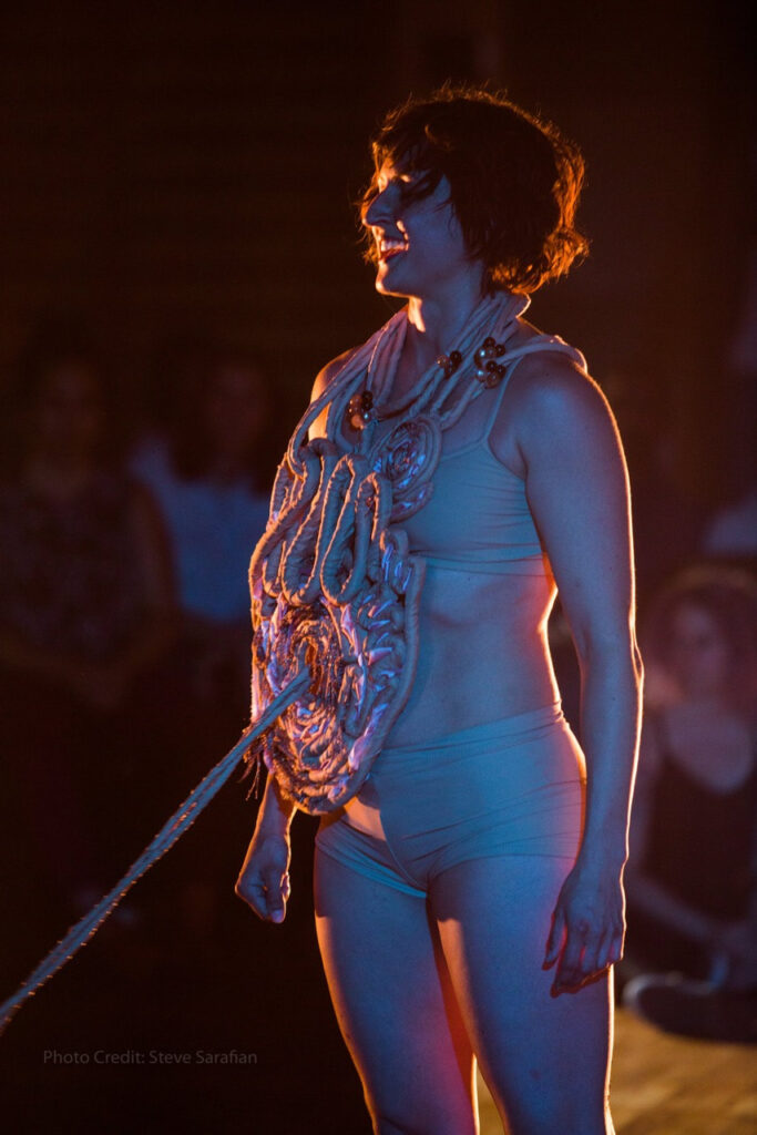 A person in a bikini standing on a stage with a rope of intestines around their neck. One rope comes from the center of their stomach out of frame.